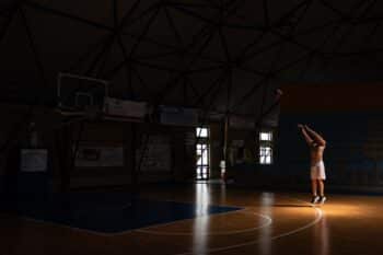 A man practices basketball shots in a dimly lit indoor court, creating a dramatic and focused atmosphere.