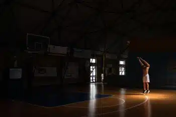 A man practices basketball shots in a dimly lit indoor court, creating a dramatic and focused atmosphere.