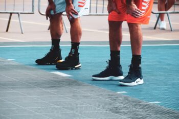 Two basketball players rest with their hands on their knees on an outdoor court, showing their sneakers and ankles