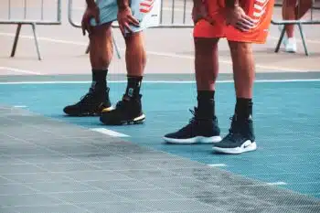 Two basketball players rest with their hands on their knees on an outdoor court, showing their sneakers and ankles
