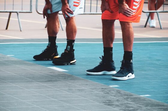 Two basketball players rest with their hands on their knees on an outdoor court, showing their sneakers and ankles