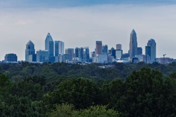 Charlotte's uptown skyline rising above a dense band of green trees under cloudy skies