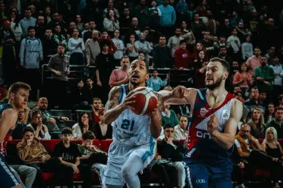 A basketball player drives toward the basket against a defender in front of a packed arena crowd